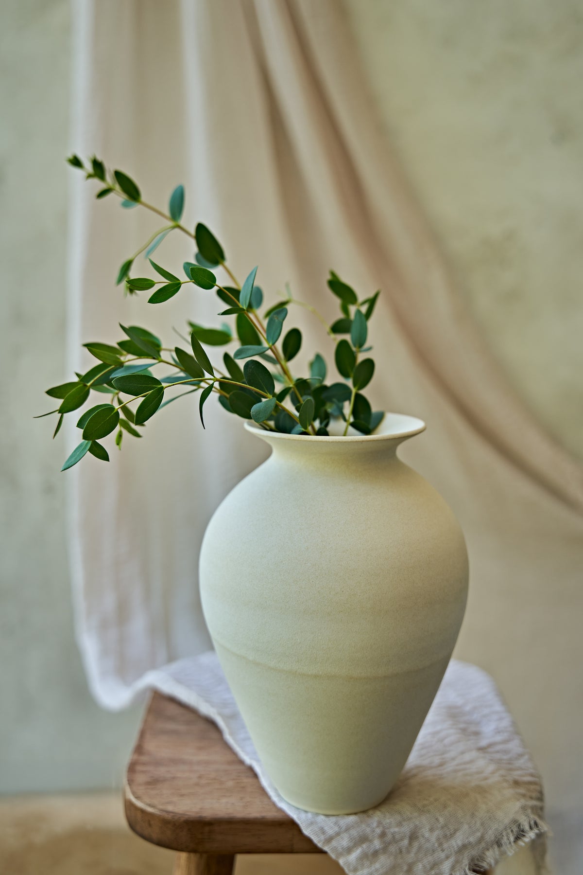A large matte ceramic vase in a warm off-white or cream color, styled with green eucalyptus branches on a small wooden stool draped with a textured linen cloth against a soft beige background.