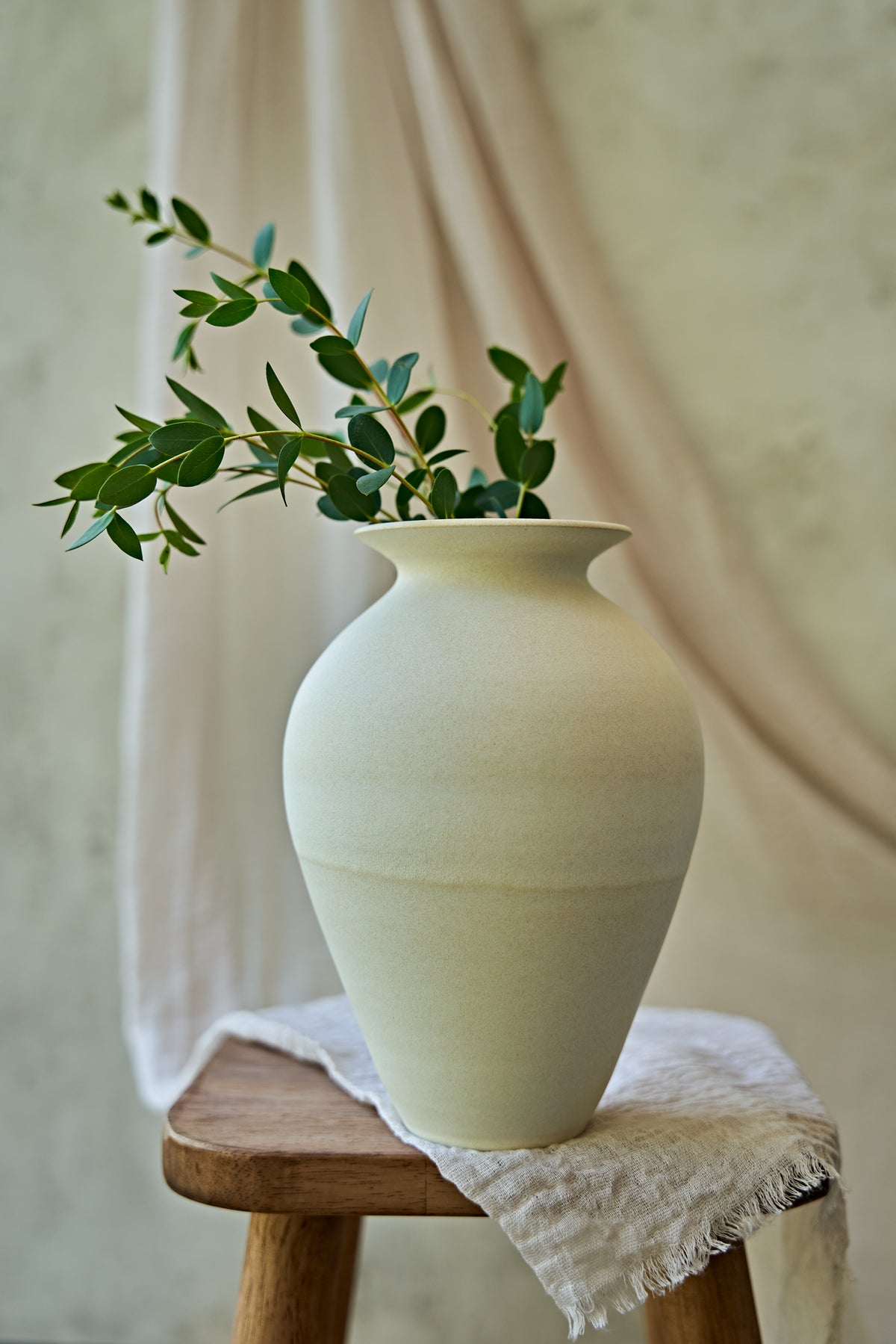 A large matte ceramic vase in a warm off-white or cream color, styled with green eucalyptus branches on a small wooden stool draped with a textured linen cloth against a soft beige background.