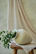 A large matte ceramic vase in a warm off-white or cream color, styled with green eucalyptus branches and lying on its side on a small wooden stool draped with a textured linen cloth against a soft beige background.