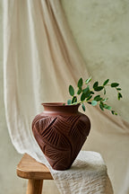 A terracotta-colored ceramic vase with carved leaf patterns sits on a wooden stool draped with cream-colored fabric, holding a green leafy sprig against a neutral background.